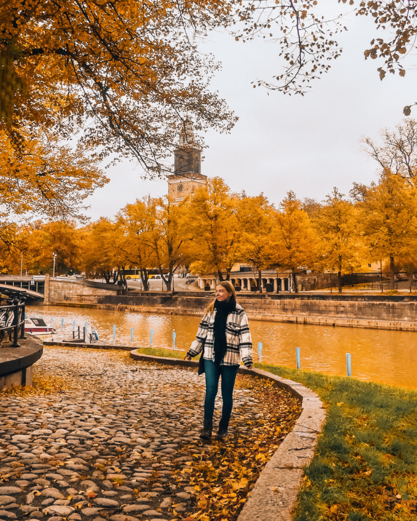 Person standing by the Aura River in Turku, with Turku Cathedral and colorful autumn trees in the background.