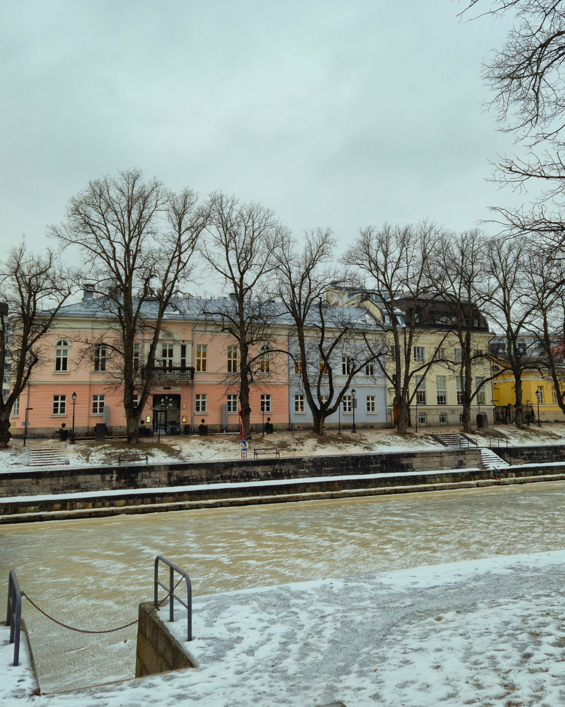 Winter view of the Aura River in Turku, Finland, with colorful riverside buildings and patches of ice on the water.