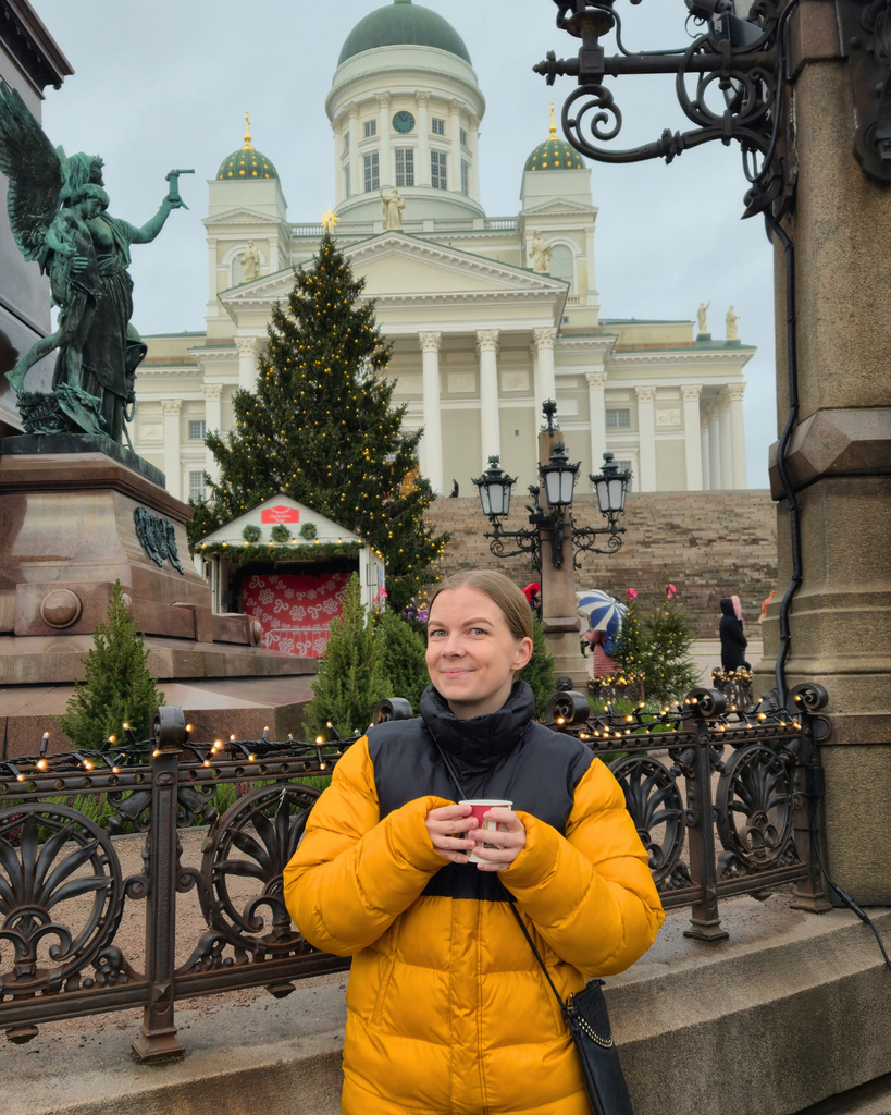A person smiling at the camera while holding a cup of glögi at Helsinki Christmas Market, with Helsinki Cathedral and festive Christmas decorations in the background.