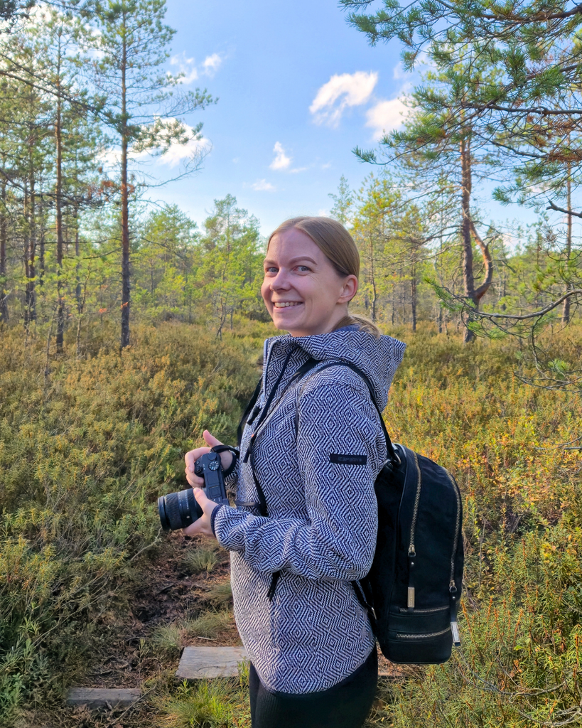 Woman smiling at the camera while holding a camera during a hike in Kurjenrahka National Park, Finland.